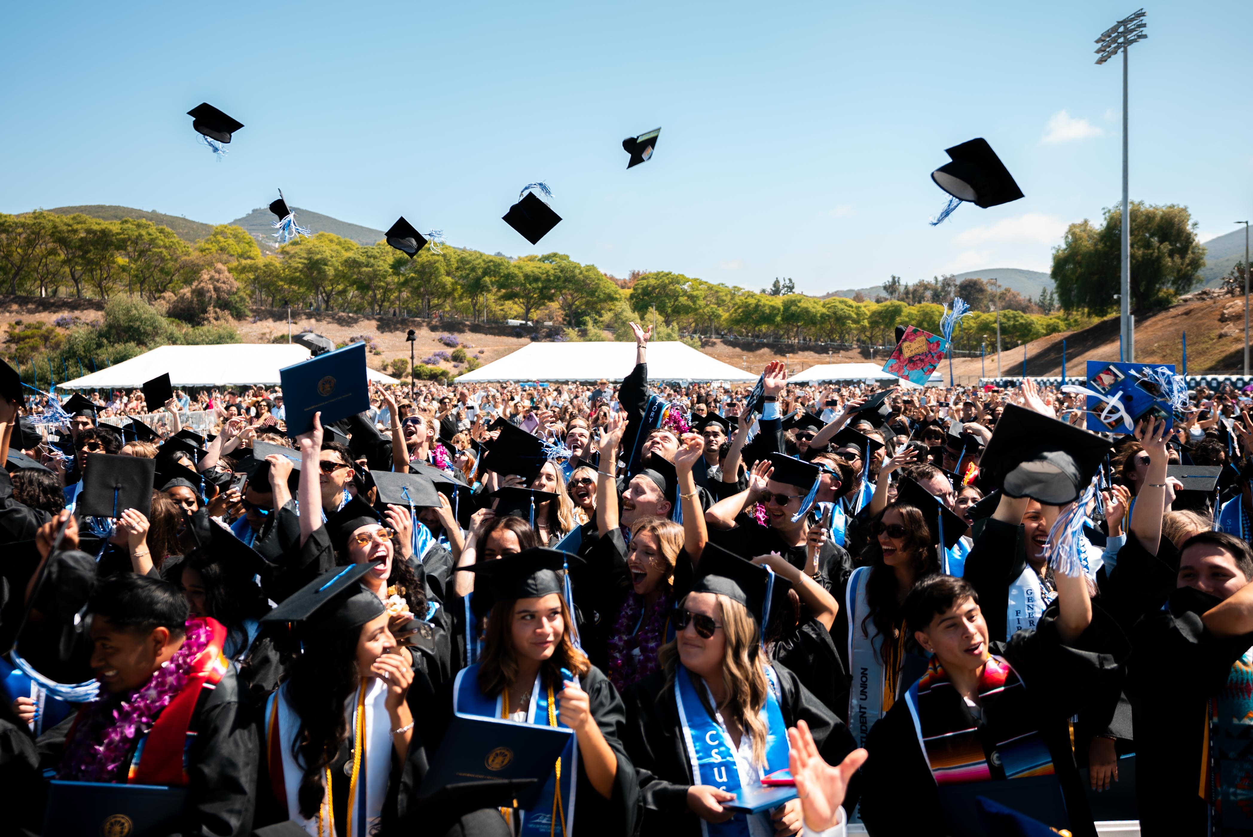 students at outdoor graduation ceremony wearing robes and caps and cheering