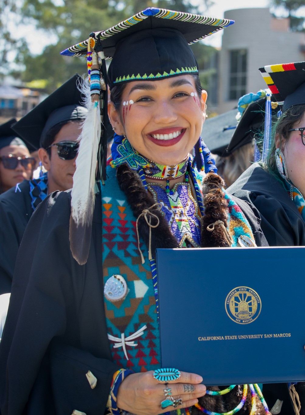Native American student wearing graduation robe and cap with traditional Native American necklace and sash